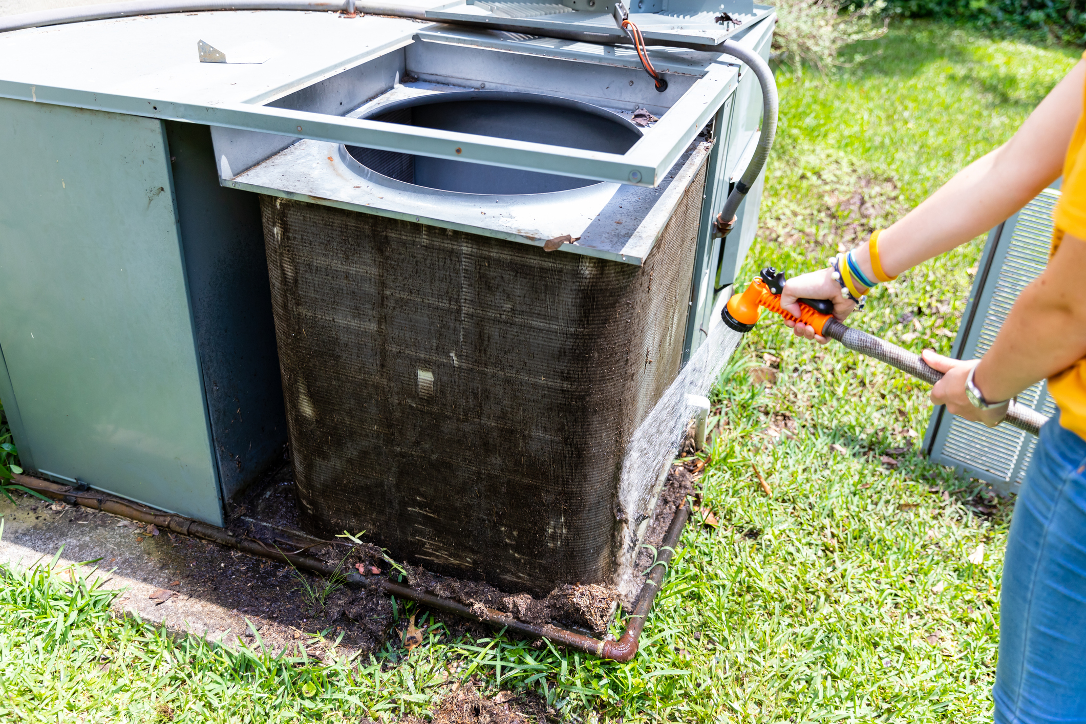 A person aiming an orange hose at a dirty outdoor central AC condenser.