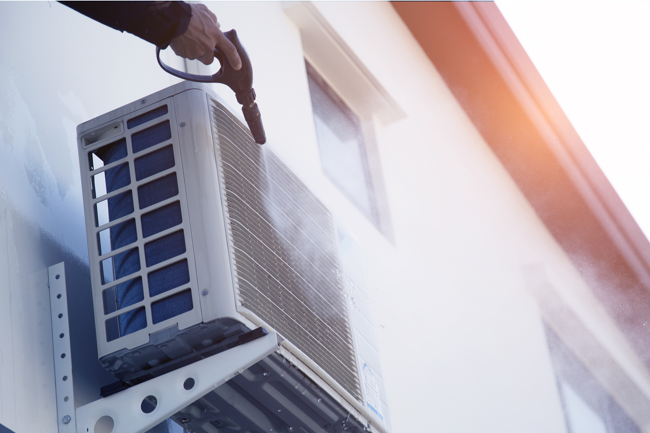 Someone's hand painting a water mister on an outdoor mini-split unit installed on the side of a white stucco home.